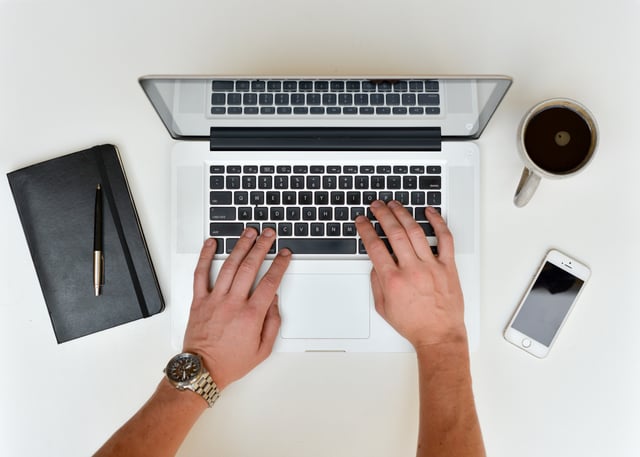 Man typing on laptop with notepad and cup of coffee on table